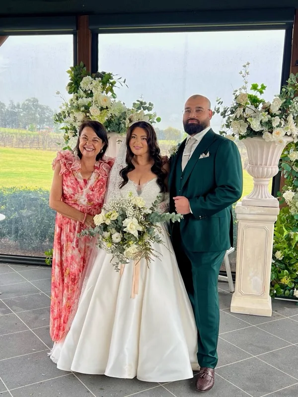 Angela with a couple beside indoor white floral arrangements.