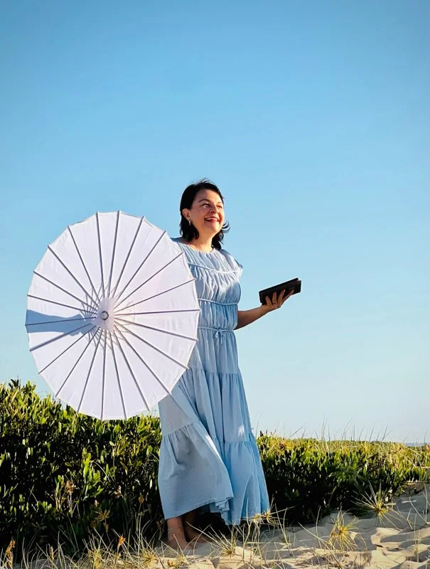 Angela holding a white parasol and ceremony book by the coast.