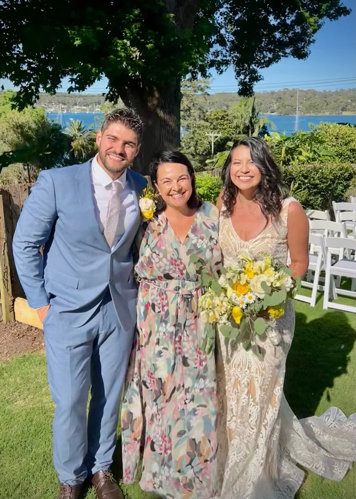 Angela with a wedding couple under a tree by the water, the bride holding yellow flowers.