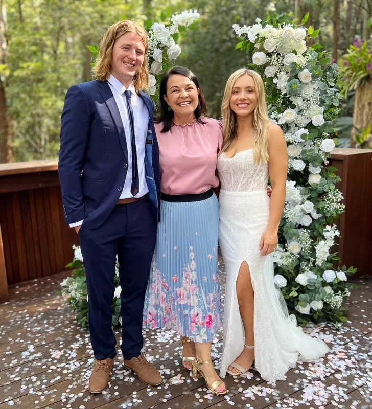 Angela in pink with a wedding couple beside a white flower arch.