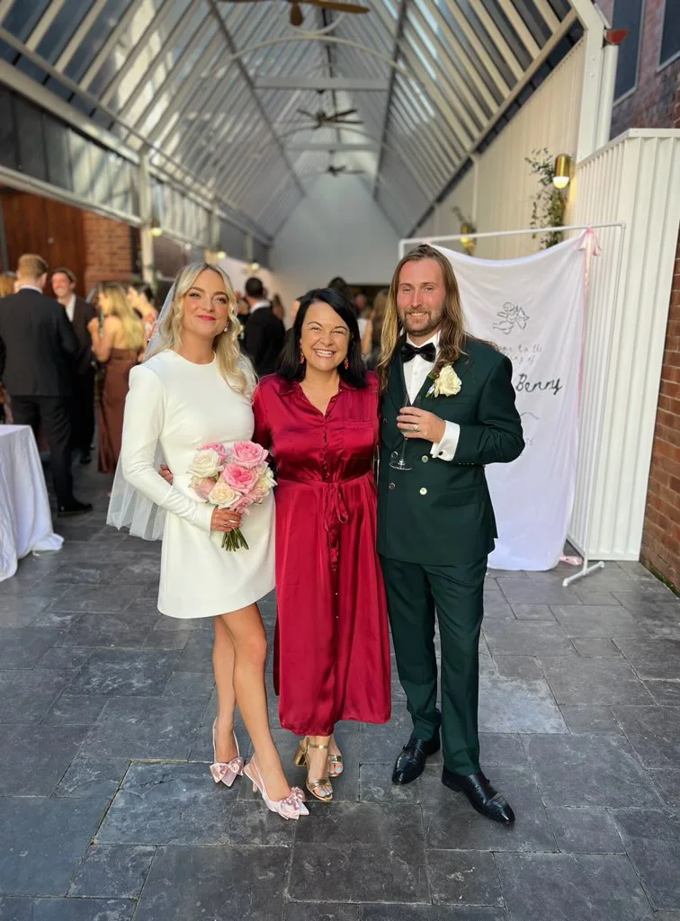 Angela in red with a wedding couple in a glass-roofed hall.