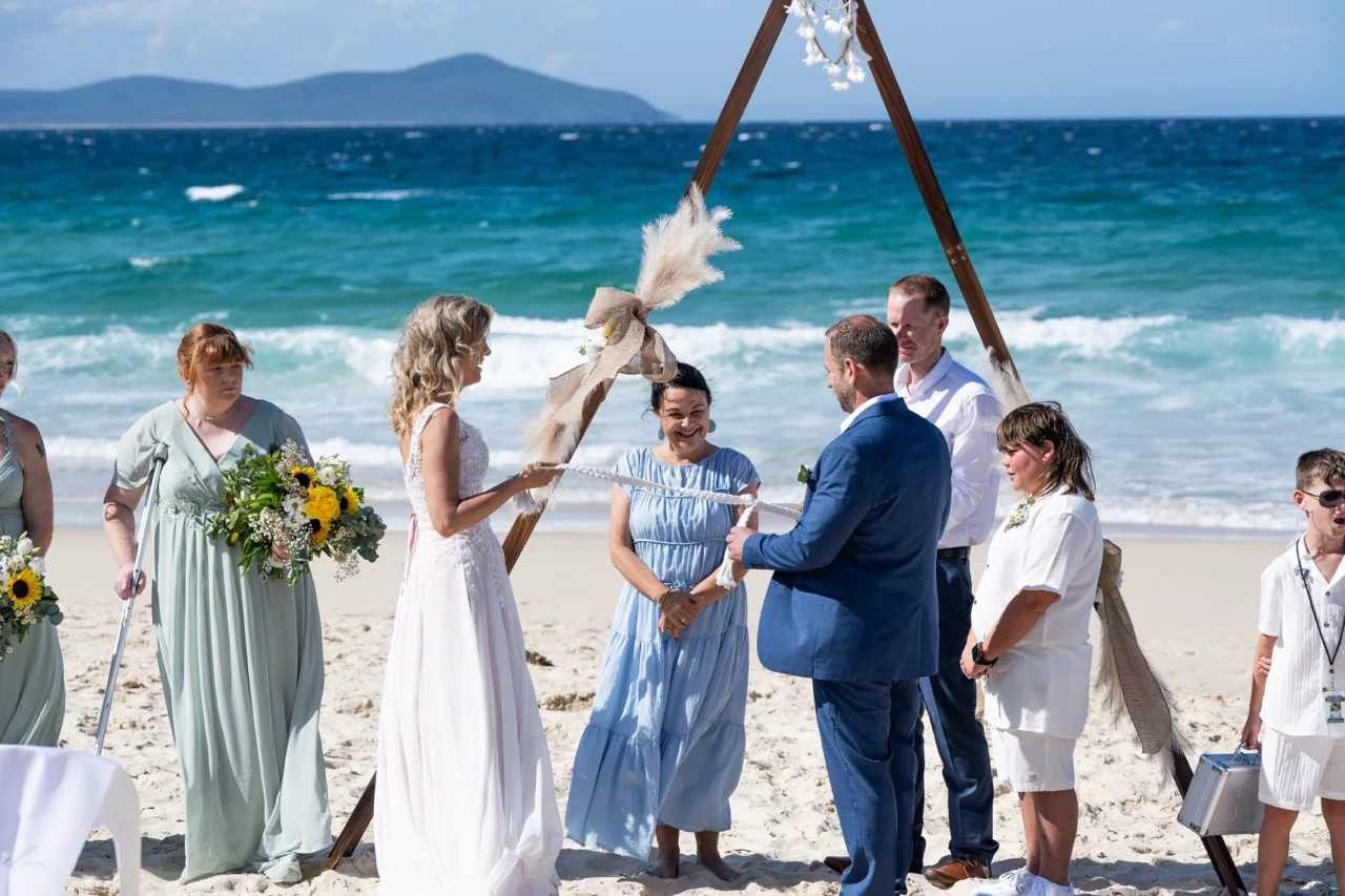 A beach ceremony under a timber arbour, with Angela beside the couple.