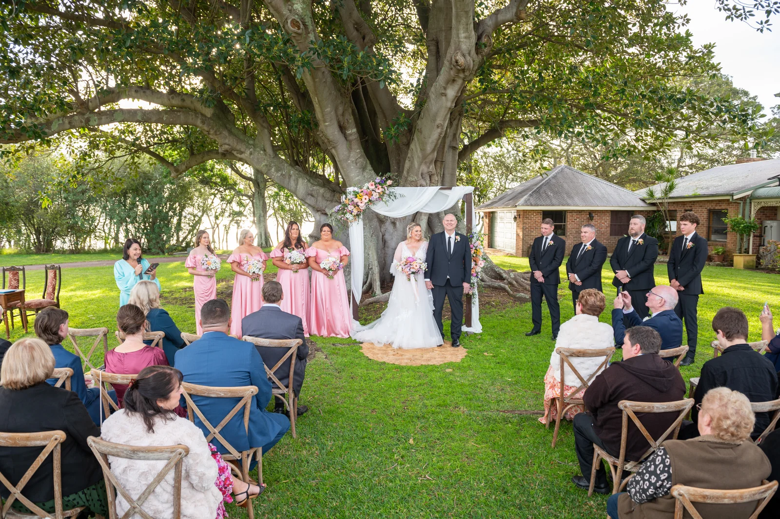 A wedding ceremony beneath a large garden tree, with guests watching the couple.